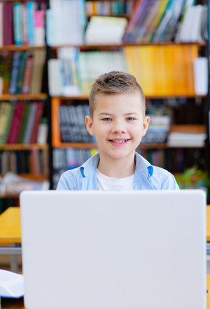 A boy with a blue shirt is working on a laptop in the library smiling on top of the laptop while...