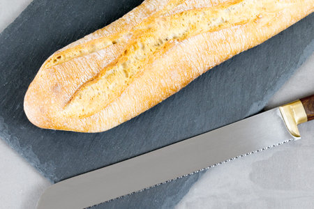 french baguette bread with knife on black cutting board on grey stone background. Diagonal.