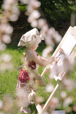 Little girl in nature paints on an easel under a blooming cherryの写真素材