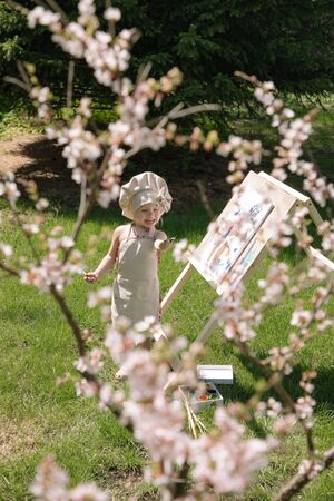 Little girl in nature paints on an easel under a blooming cherryの写真素材