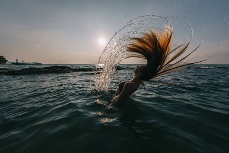 Girl with long hair swims in the sea, splashes water on a background of a sunset skyの写真素材