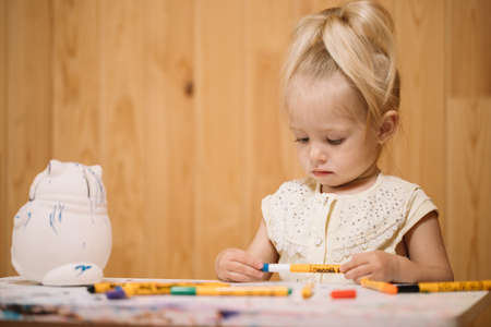Little girl paints a clay piggy bank in the form of a fat cat with color markers on a wooden wall backgroundの写真素材