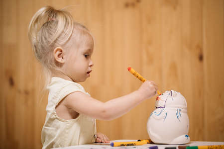 Little girl paints a clay piggy bank in the form of a fat cat with color markers on a wooden wall backgroundの写真素材
