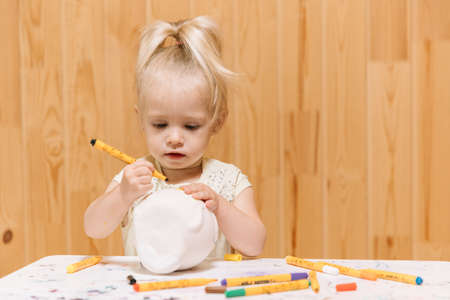 Little girl paints a clay piggy bank in the form of a fat cat with color markers on a wooden wall backgroundの写真素材