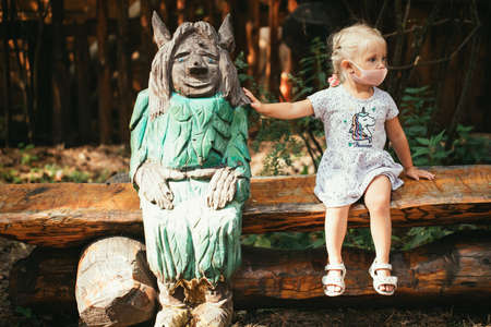 Little girl in a medical mask sits on a bench in a city park near a wooden figureの写真素材