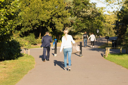 Different Passers-by Walk Along the Asphalt Path of Street Among Green Trees on a Sunny Day. English...