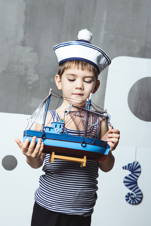 Cute cabin boy in  striped t-shirt and sailor's caps holding a model of the shipの写真素材