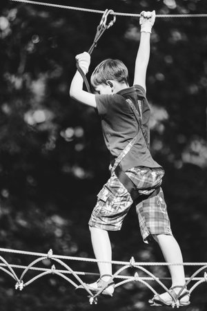 Boy passing cable route in extreme adventure rope park on sunny summer day, black and white shot