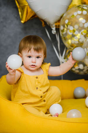 1 year birthday baby girl playing with ball pool balls with festive balloons on background, yellow and gray colorsの写真素材