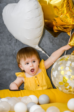 1 year birthday baby girl playing with ball pool balls with festive balloons on background, yellow and gray colorsの写真素材