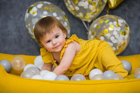1 year birthday baby girl playing with ball pool balls with festive balloons on background, yellow and gray colorsの写真素材
