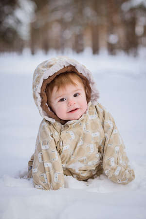 Cute one year old baby girl sitting in deep snow in forest, beige overallの写真素材