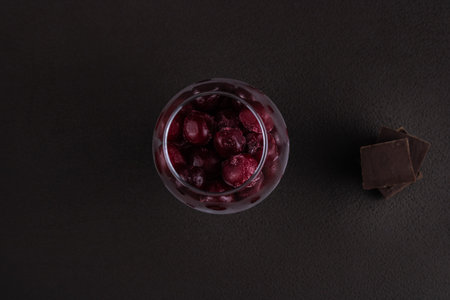 Glass glass with frozen cherries and chocolate on a dark brown background, top view.