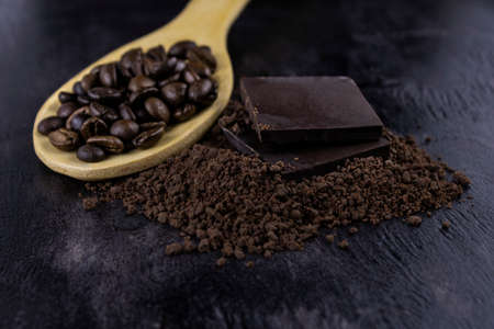 a crumb of dark chocolate and two whole bars with coffee beans on a wooden spoon on a dark background close-upの写真素材