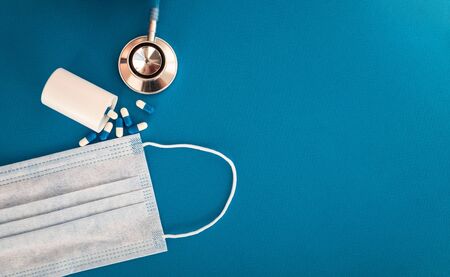 Composition with medical stethoscope, disposable face masks, overdropped plastic container of pills on a blue background. Top view. Free copy spaceの写真素材