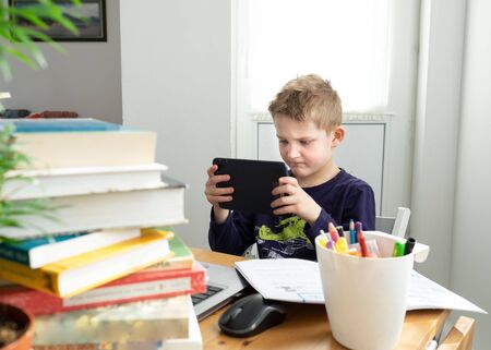 Caucasian preteen boy play game on tablet instead of doing distance learning task. Books and copybooks around. Concept of distance learning in isolationの写真素材