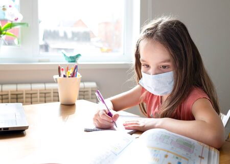 Caucasian preteen girl with medical mask on her face concentrated on her task with textbook. Concept of distance learning in isolation while coronavirusの写真素材