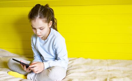 Concept of distance education, free time, technology and internet. Preteen caucasian little student girl with tablet pc at home sitting on couch on yellow backgroundの写真素材
