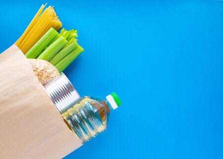 Coronavirus food supplies concept. Food layout with pasta, sunflower oil, dried peas, canned food, celery in paper bag on blue background. Flat lay. Copy spaceの写真素材