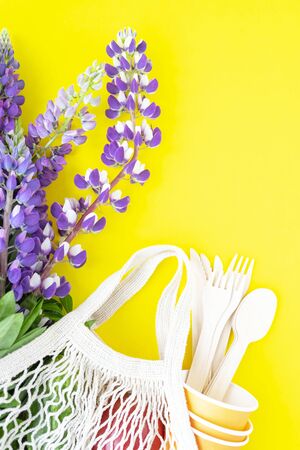 A string bag with a bouquet of fresh purple lupins, cardboard disposable cups and wooden spoons, forks, knives on a yellow background. The concept of zero waste and no plasticの写真素材