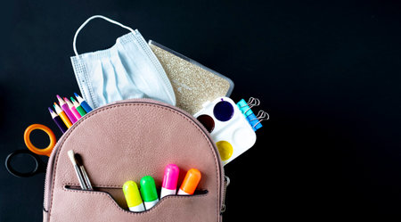 Concept back to school. Full beige School Backpack with supplies and medical mask on black chalkboard background. Flat lay.の写真素材