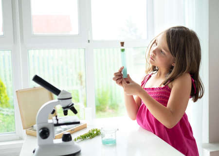 Pretty blonde little girl working with test tube and microscope. The kid learning science in the school laboratory or at home. Child looking make experiment with microscope on the table.の写真素材
