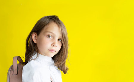 serious pretty schoolgirl. cute caucasian girl with blond hair with backpack on her back on yellow...