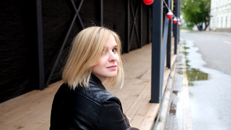 Smiling pretty blond young woman in black leather jacket and red shirt sitting outdoor on the street...