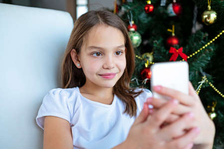 Close up smiling caucasian young preteen girl talking on phone, sitting near festive Christmas tree at home, happy female holding smartphone, chatting with friends or relatives, congratulatingの写真素材