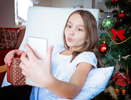 Close up smiling caucasian young preteen girl talking on phone, sitting near festive Christmas tree...