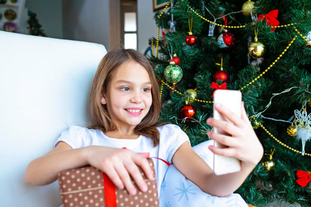 Close up smiling caucasian young preteen girl talking on phone, sitting near festive Christmas tree...