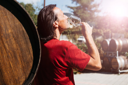 A handsome adult man stands near a wine barrel on a farm and tastes a glass of white wine. Sunny...