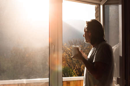An adult man with a mug of hot tea stands and looks at the mountains. The view from the window of the house.の写真素材