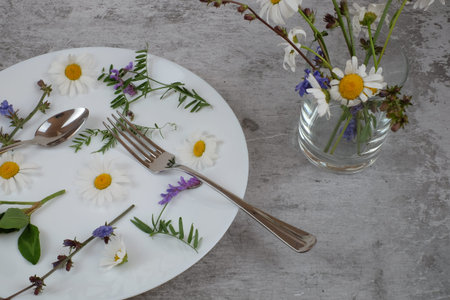 Flowers in white plate with fork and spoon on grey textured background, vase with wilf flowers,...
