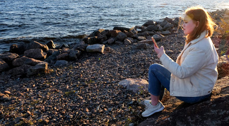 Girl watching smartphone on rock seaside background, lifestyle banner with copy space, warm sun...
