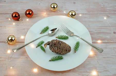 Christmas diet concept. Pine cones and pine branches as food on whete plate, fork and knife on wooden table, lights and balls decoration. Vegan menu concept.の写真素材