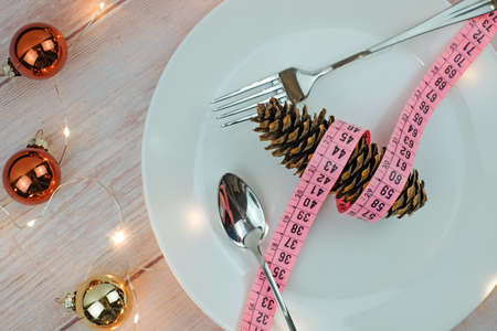 Christmas diet concept. Pine cones with tape measure on white plate, fork and spoon on wooden table, lights and balls decoration. Vegan menu concept.の写真素材
