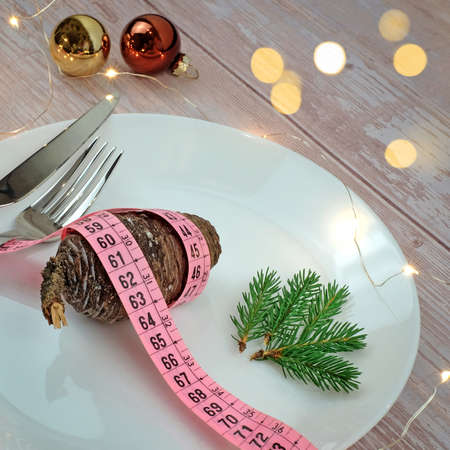 Christmas diet concept. Pine cones with tape measure on white plate, fork and knife on wooden table. Golden lights bokeh and balls decoration. Vegan menu concept. Square image.の写真素材
