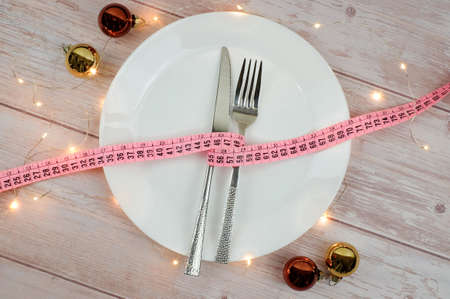 Diet concept for christmas dinner. The tape measure with fork and knife on white plate, on wooden table. Lights, bokeh and balls decoration. Table setting. Top view.の写真素材