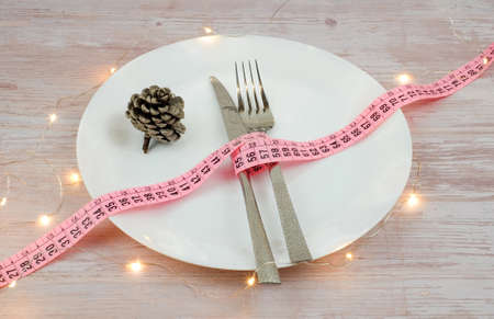 Diet concept for christmas dinner. The tape measure with fork and knife on white plate and pine cone on wooden table. Lights decoration.の写真素材