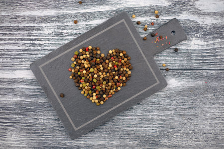 Colored peppercorns heart shape on the gray stone cutting board on gray wooden background, selective...