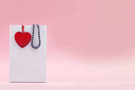 White gift bag with romantic red heart and pearl beads on pink background. Valentines holiday sale. Copy space, mock-upの写真素材