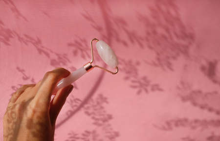 Face roller made of rose quartz in woman hand on pink background with openwork shadows. Anti-aging therapy, alternative medicine. Copy space.の写真素材