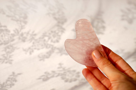 Face scraper made of rose quartz in woman hand on white abstract background. Anti-aging therapy,...