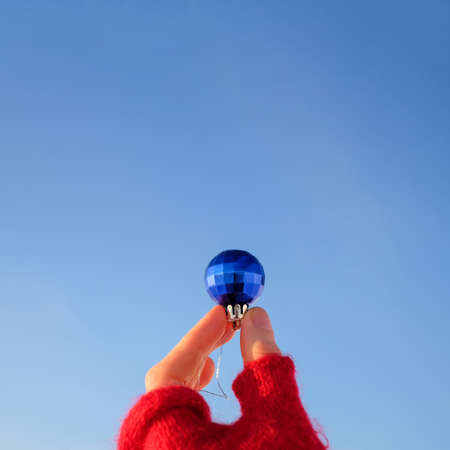 Hand in red glove holding Christmas ball on blue clear sky background, square photo with copy spaceの写真素材