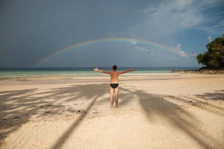 A man stands on Loh Moo Dee Beach, against the background of the sea and the rainbow. Thailand, Phi Phi island.の写真素材