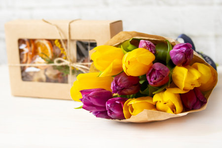 Fresh bouquet purple yellow tulips in kraft packaging with a box of handmade sweets. Candy and flowers, a gift for the woman he lovesの写真素材