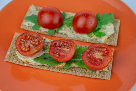 Rye crisps with cherry tomatoes, arugula and hummus on a bright orange plate. Delicious healthy vegan snack.の写真素材
