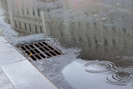 Autumn asphalt, metal grating stormwater on a city street. Autumn rainy weather with puddles, outflow of water from the highway. Reflection of urban building in a puddle, backdrop texture backgroundの写真素材