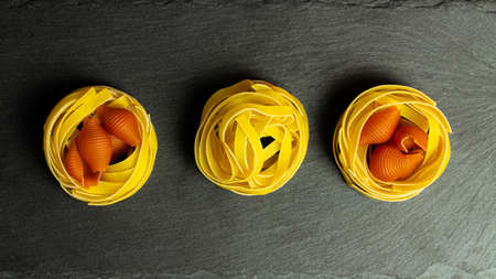 Three bunches of pasta fettuccine on a black background, raw yellow egg pasta nest with red shells concilia from the dough. Italian traditional food, still lifeの写真素材